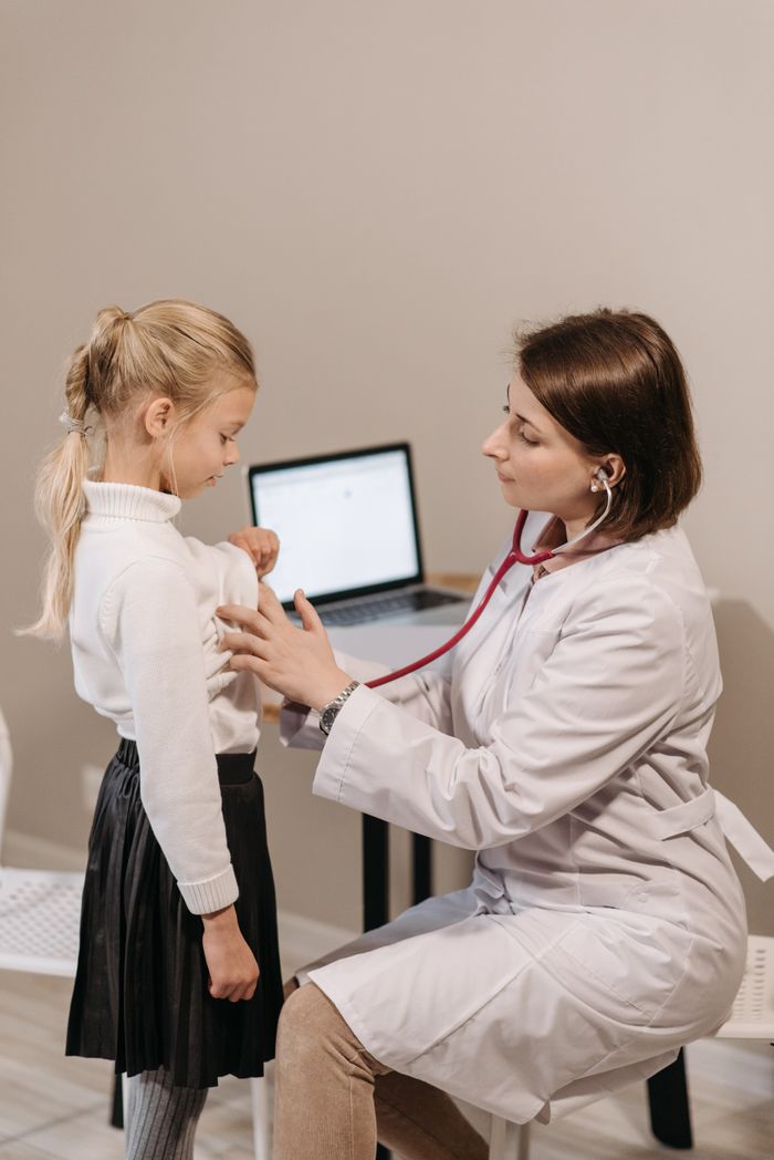 Home A female doctor conducts an examination on a young girl with a stethoscope in a clinic setting.