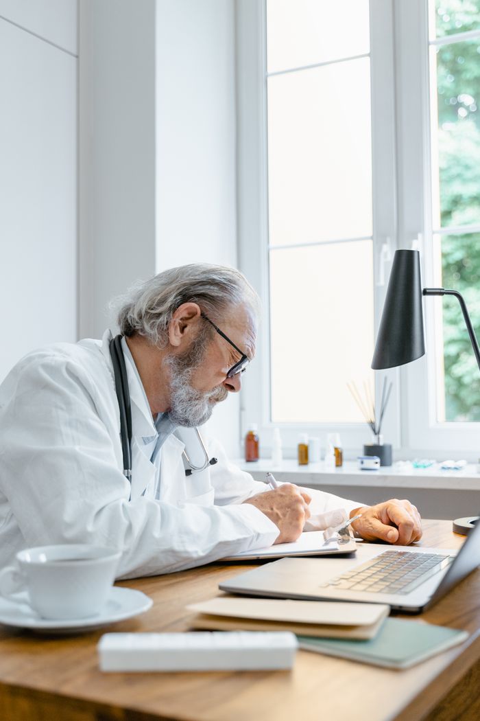 Senior male doctor writing notes during a telehealth session in a bright office.